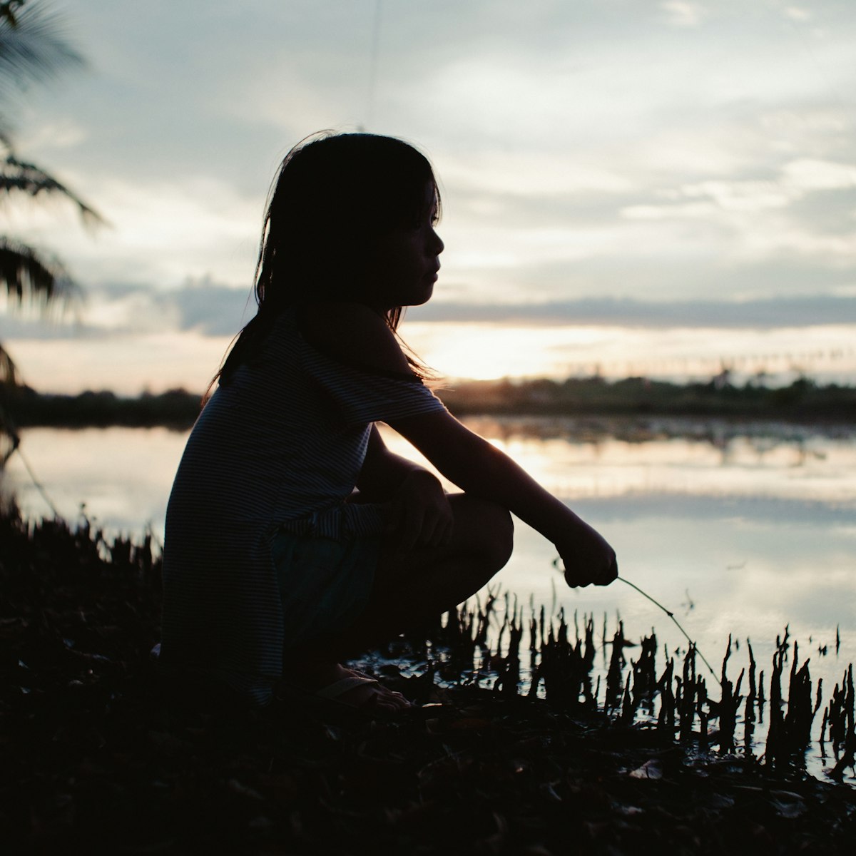A child crouched at the river's edge at dusk, silhouetted against still water.